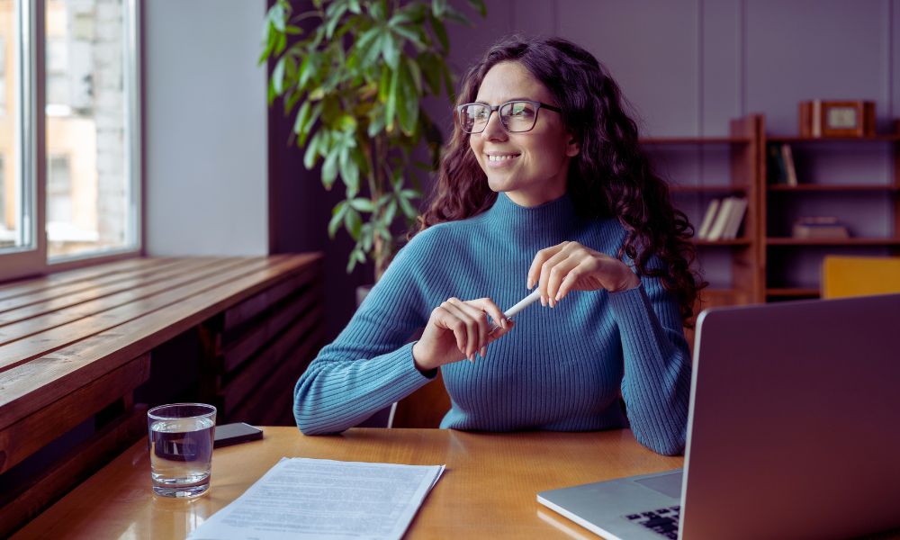 A woman with long curly hair and glasses sits at a desk with papers, a laptop, and a glass of water, smiling and looking out the window in a bright office setting where Humanico helps foster productivity. | Humanico Workforce Intelligence Software
