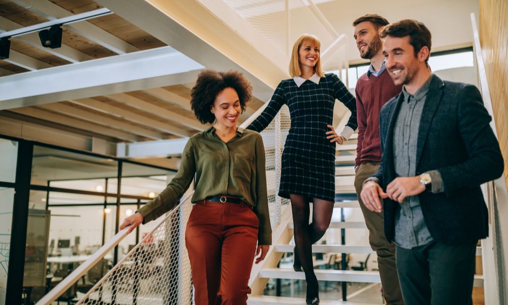Four professionally dressed people, two men and two women, are smiling and walking down an office staircase together in a modern, bright workspace with glass walls and exposed ceilings—showcasing how Humanico helps teams connect. | Humanico Workforce Intelligence Software