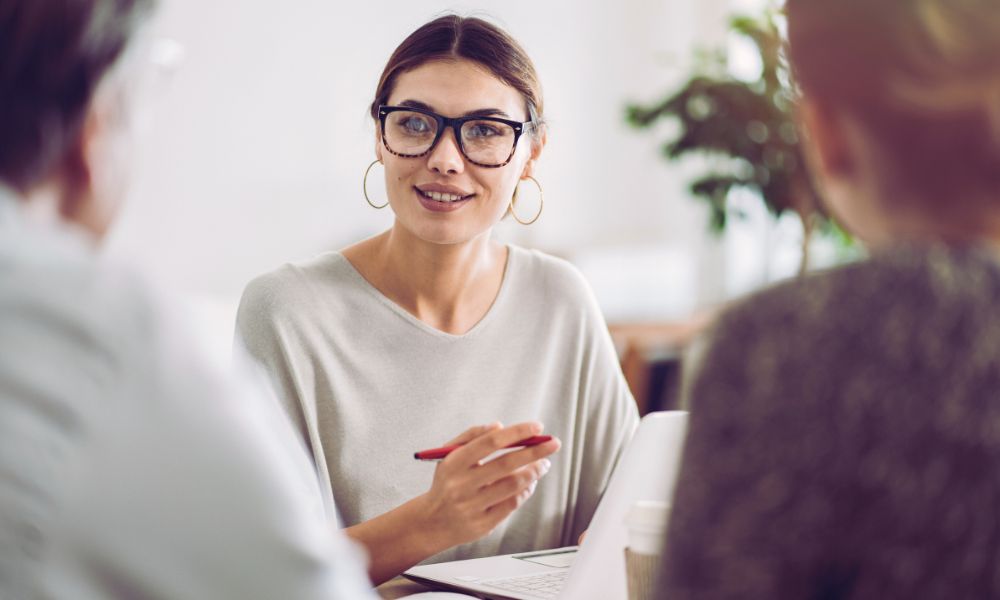 A woman wearing glasses and hoop earrings sits at a table with two others, holding a pen and smiling, engaged in conversation. A laptop and coffee cup are visible in front of her as Humanico helps foster collaboration. | Humanico Workforce Intelligence Software