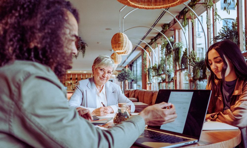 At a bright, plant-filled cafe, three people sit at a table. One person points to a laptop screen displaying a document as another listens and an older woman in glasses smiles, observing how Humanico helps foster collaboration. | Humanico Workforce Intelligence Software
