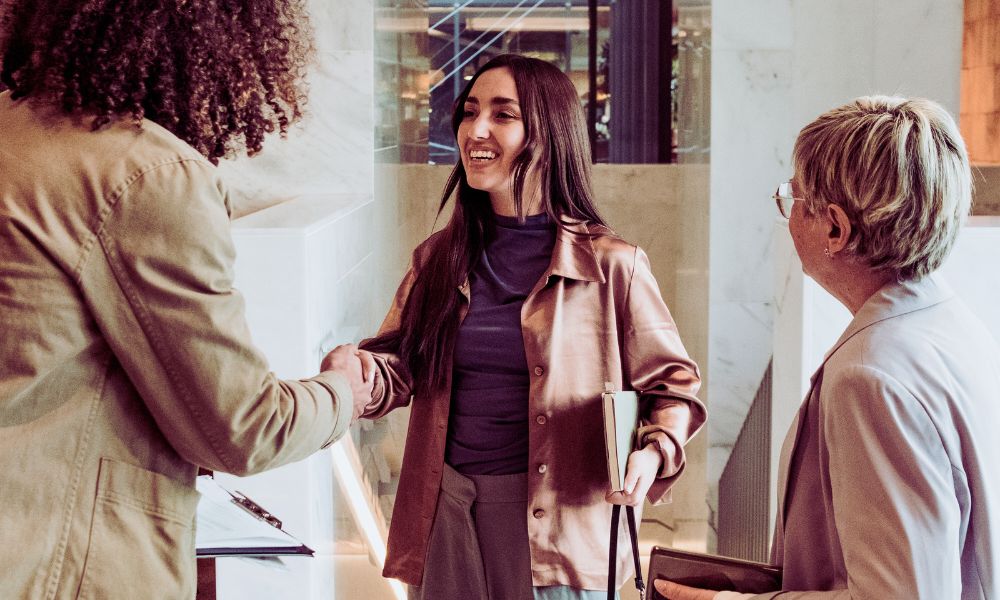 Three people stand indoors; a young woman in a satin shirt smiles and shakes hands with someone with curly hair, as an older person with glasses observes, holding a handbag. Humanico helps foster these positive connections. | Humanico Workforce Intelligence Software