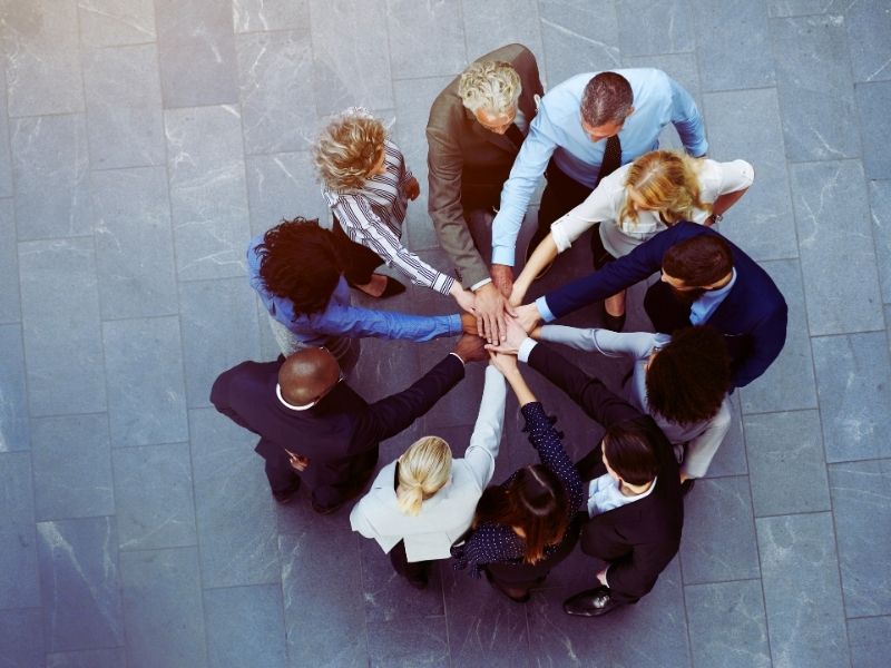 A group of diverse people in business attire stand in a circle on a tiled floor, reaching into the center to stack their hands together in a gesture of teamwork and unity—symbolizing stronger teams working together from above. | Humanico Workforce Intelligence Software