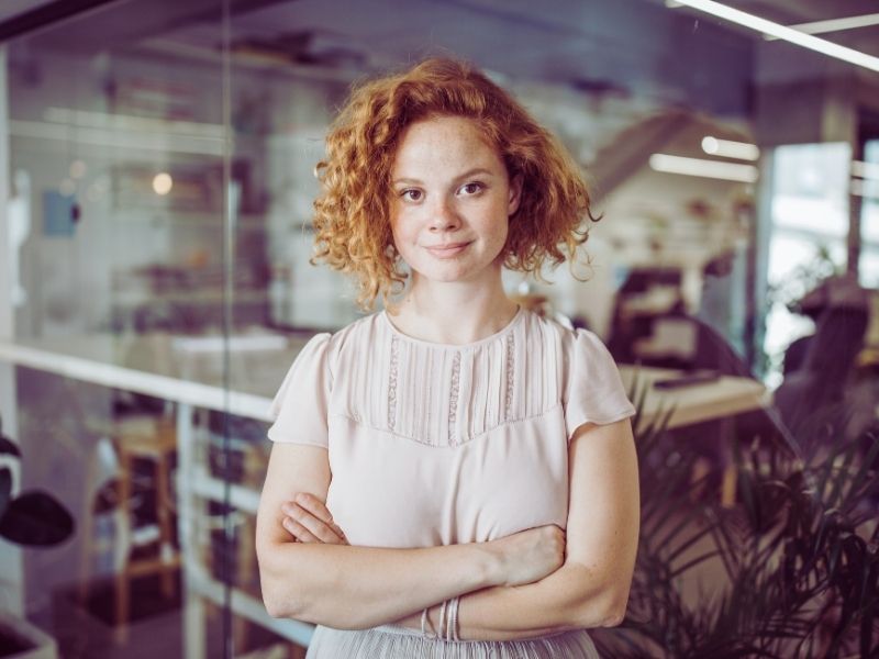 A woman with curly red hair, wearing a light pink blouse, stands with arms crossed and smiles confidently in a modern office—her hidden talent shining through amid the glass walls and shelves in the background. | Humanico Workforce Intelligence Software