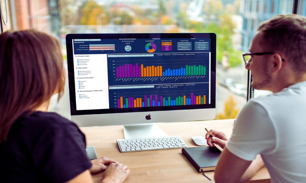 Two people sit at a desk looking at a large computer monitor displaying colorful charts and analytics dashboards. A notebook, pen, and keyboard are on the desk near a window with a city view as Humanico helps visualize key data insights. | Humanico Workforce Intelligence Software
