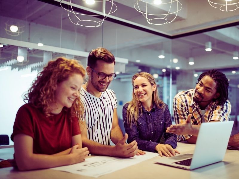 Four young adults sit at a table in a modern office, smiling and talking while looking at papers and a laptop. One gestures toward the computer, sparking a collaborative discussion about how to Measure What Matters in their latest project. | Humanico Workforce Intelligence Software
