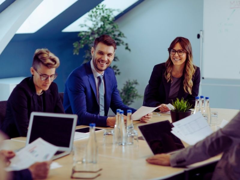 Four people in business attire sit around a conference table with laptops, papers, and water bottles, smiling and engaged in discussion as they navigate change in a modern office setting. | Humanico Workforce Intelligence Software