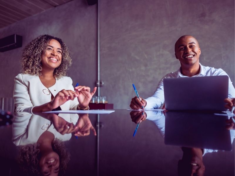 Two people sit at a glossy conference table smiling; one holds a pen and paper, the other has a laptop. They appear to be engaged in leadership planning in a modern, well-lit office space. | Humanico Workforce Intelligence Software