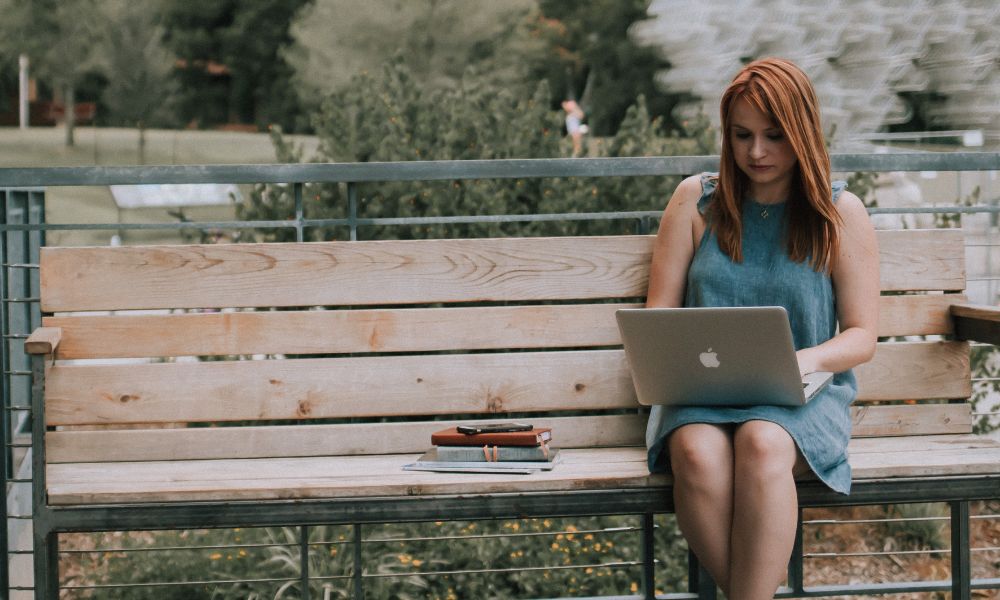 A woman with long red hair sits on a wooden bench outdoors, working on a laptop. Books and notebooks are beside her as she researches employee retention analytics. Trees and greenery fill the background. | Humanico Workforce Intelligence Software