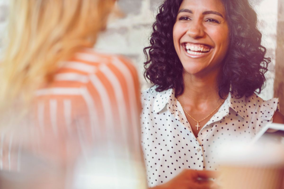 A woman with curly hair wearing a white polka dot blouse and a cross necklace smiles brightly while discussing hr tools for new managers with another person in a casual setting. | Humanico Workforce Intelligence Software