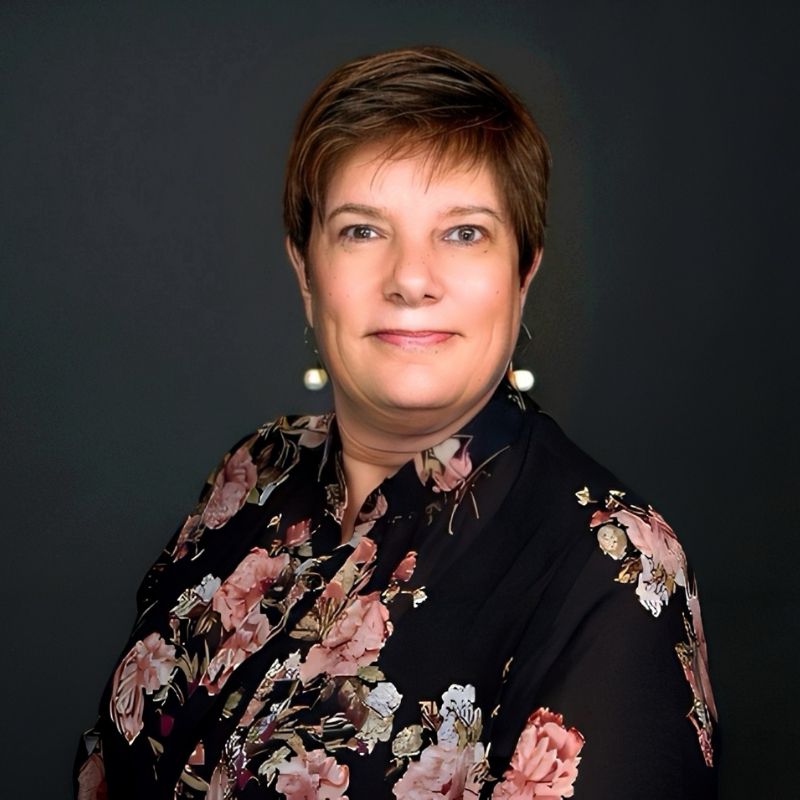 Juliette Connolly, a woman with short brown hair in a black floral blouse, poses in front of a dark background, smiling gently at the camera. She has light skin and is wearing drop earrings. | Humanico Workforce Intelligence Software