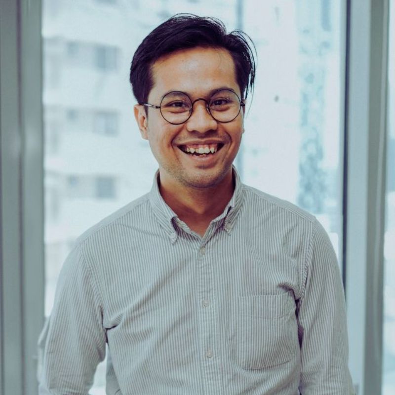 A man with glasses and short dark hair, wearing a light gray striped button-up shirt, stands smiling in front of large windows with a blurred cityscape in the background, representing the effectiveness of HR tools for executives. | Humanico Workforce Intelligence Software