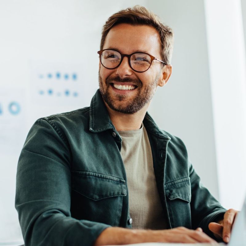A smiling man with short brown hair, a beard, and glasses, wearing a green jacket over a beige shirt, sits indoors with a laptop—perhaps exploring HR software Australia recommends as top tools for new managers. | Humanico Workforce Intelligence Software