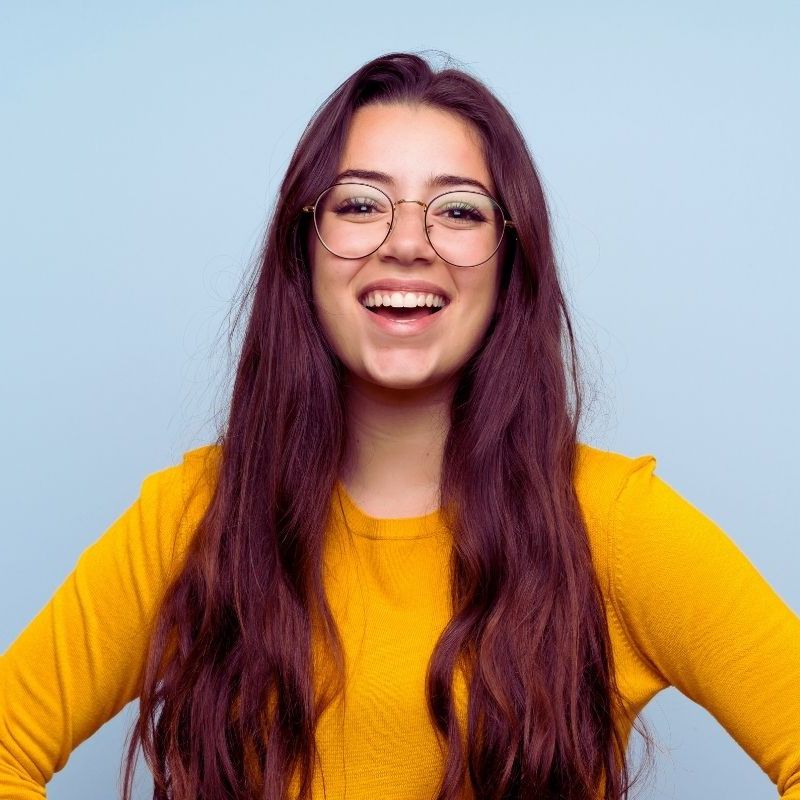 A young woman with long brown hair, wearing round glasses and a bright yellow sweater, smiles confidently against a light blue background—ready to take on onboarding for new managers. | Humanico Workforce Intelligence Software