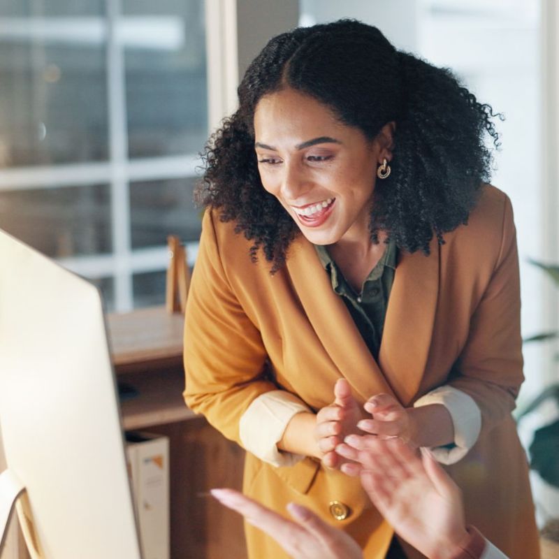 A woman in a brown blazer smiles and claps her hands while looking at a computer screen, celebrating a successful onboarding for new managers, with another pair of hands visible in the foreground, highlighting teamwork and achievement. | Humanico Workforce Intelligence Software
