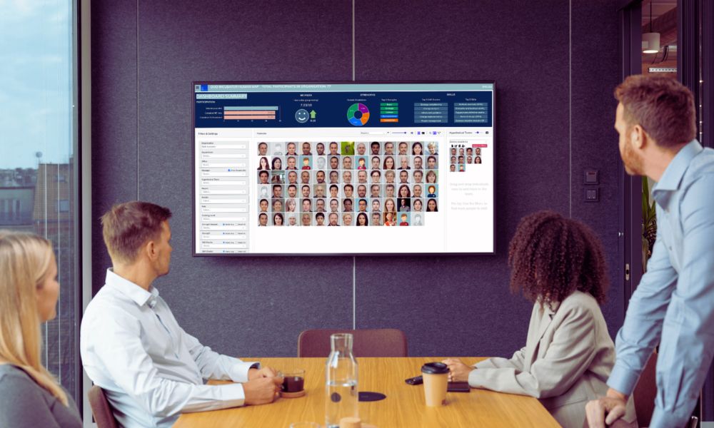 Three people sit at a conference table, while a fourth stands, all focused on a large wall-mounted screen displaying HR tools for executives—headshots, graphs, and charts—in a modern office meeting room. | Humanico Workforce Intelligence Software