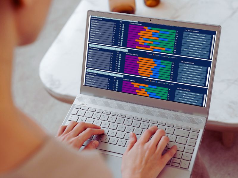 A person types on a laptop displaying colorful bar graphs and data charts, indicating analysis or comparison of information to support building stronger teams. The background is blurred, focusing on the laptop and hands. | Humanico Workforce Intelligence Software