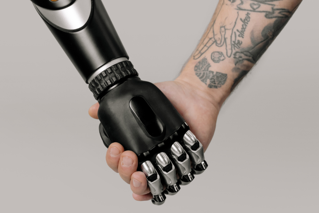 A human hand with tattoos holds the hand of a black and silver robotic prosthetic arm against a plain, light background, symbolizing the evolution of humanico—a blend of humanity and technology. | Humanico Workforce Intelligence Software